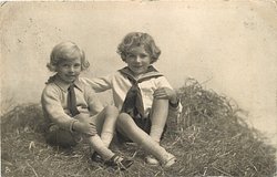 two boys play in straw, both sit cross-legged, looking forward