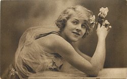 woman with fluffy hair faces right, looks up and left, rests her elbows on table, holds roses