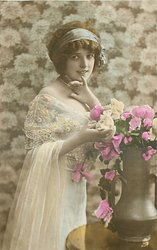 glamorous lady in dress with lace top & filmy shawl over her arm, bunch of roses in copper vase, looks at camera but body turned to right, right hand touches flowers, left hand under chin