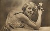 Image 1 - woman with fluffy hair faces right, looks up and left, rests her elbows on table, holds roses