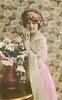 Image 1 - glamorous lady in dress with lace top & filmy shawl over her arm, bunch of roses in copper vase, looks at camera, hands pressed together in front of chest