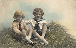 two boys play in straw, both sit cross-legged, looking forward