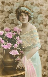 glamorous lady in dress with lace top & filmy shawl over her arm, bunch of roses in copper vase, looks at camera, left hand touches table