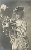 Image 1 - girl stands looking half left holding floral basket held in front of her