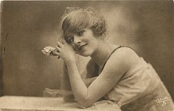 woman with fluffy hair faces left, looks up, rests her elbows on table, and rests her head on hands, one rose held in her clasped hand