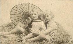 two boys play in straw, looking forward, parasol behind, handle to left of boy on left